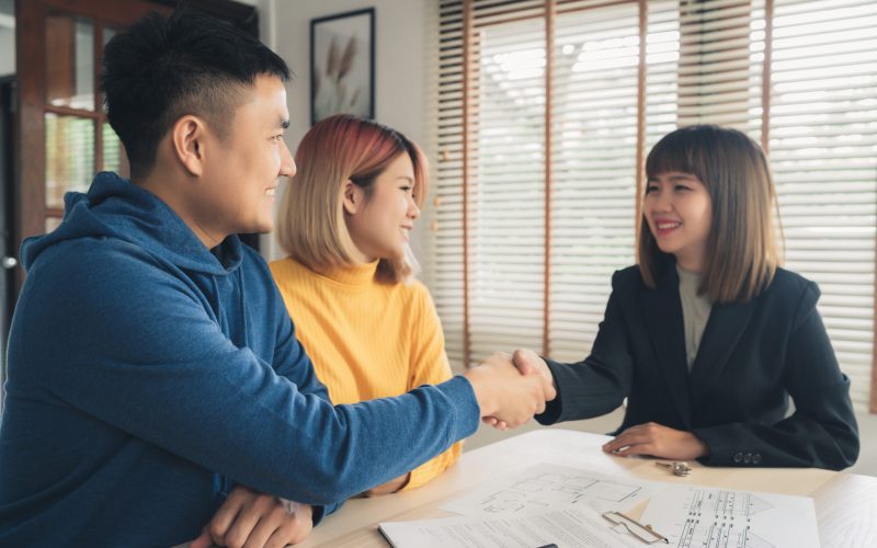 Happy young Asian couple and realtor agent. Cheerful young man signing some documents and handshaking with broker while sitting at desk. Signing good condition contract.
